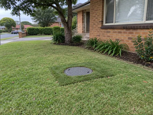 Flush sewer access cover in a front lawn beside a brick house in Kingsgrove